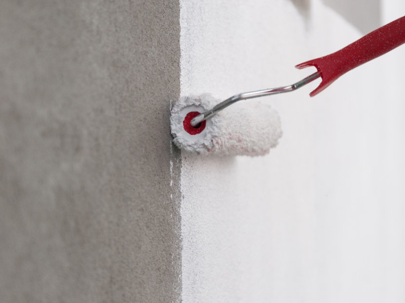 Eine Farbrolle trägt weiße Fassadenfarbe auf eine verputzte Hauswand auf und sorgt für eine gleichmäßige Beschichtung der Oberfläche.
