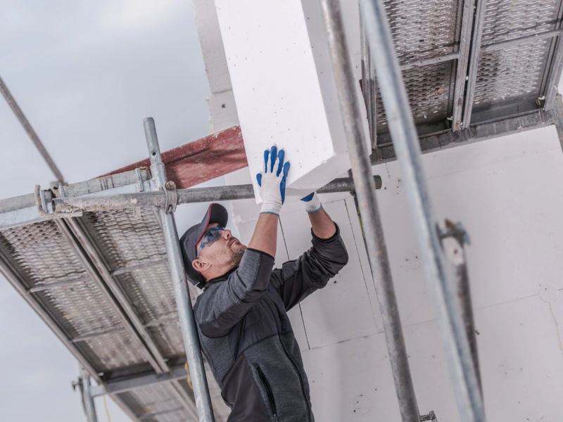 Ein Handwerker befestigt Dämmplatten an der Fassade eines Gebäudes, während er auf einem Baugerüst steht – typische Arbeit bei einer energetischen Fassadendämmung.