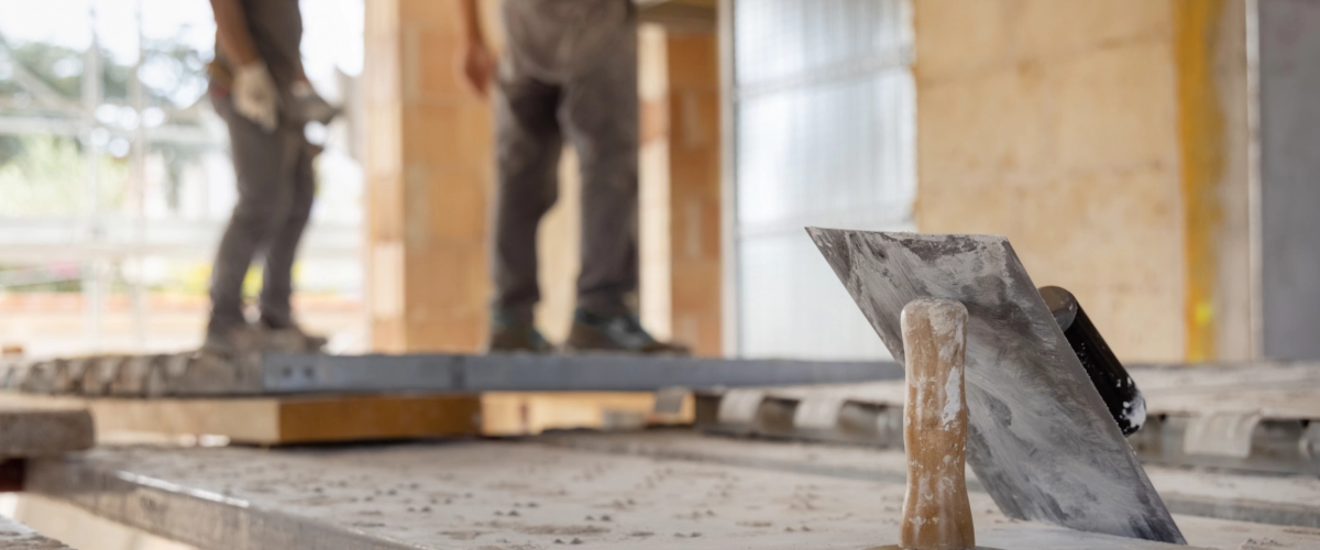Detailaufnahme einer Kelle für Putzarbeiten auf einer Baustelle, während im Hintergrund Handwerker an Bau- oder Fassadenarbeiten arbeiten.