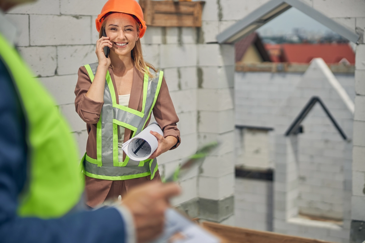 Eine Bauleiterin mit Sicherheitshelm und Bauplan koordiniert telefonisch Arbeiten auf einer Baustelle während eines Bau- oder Sanierungsprojekts.